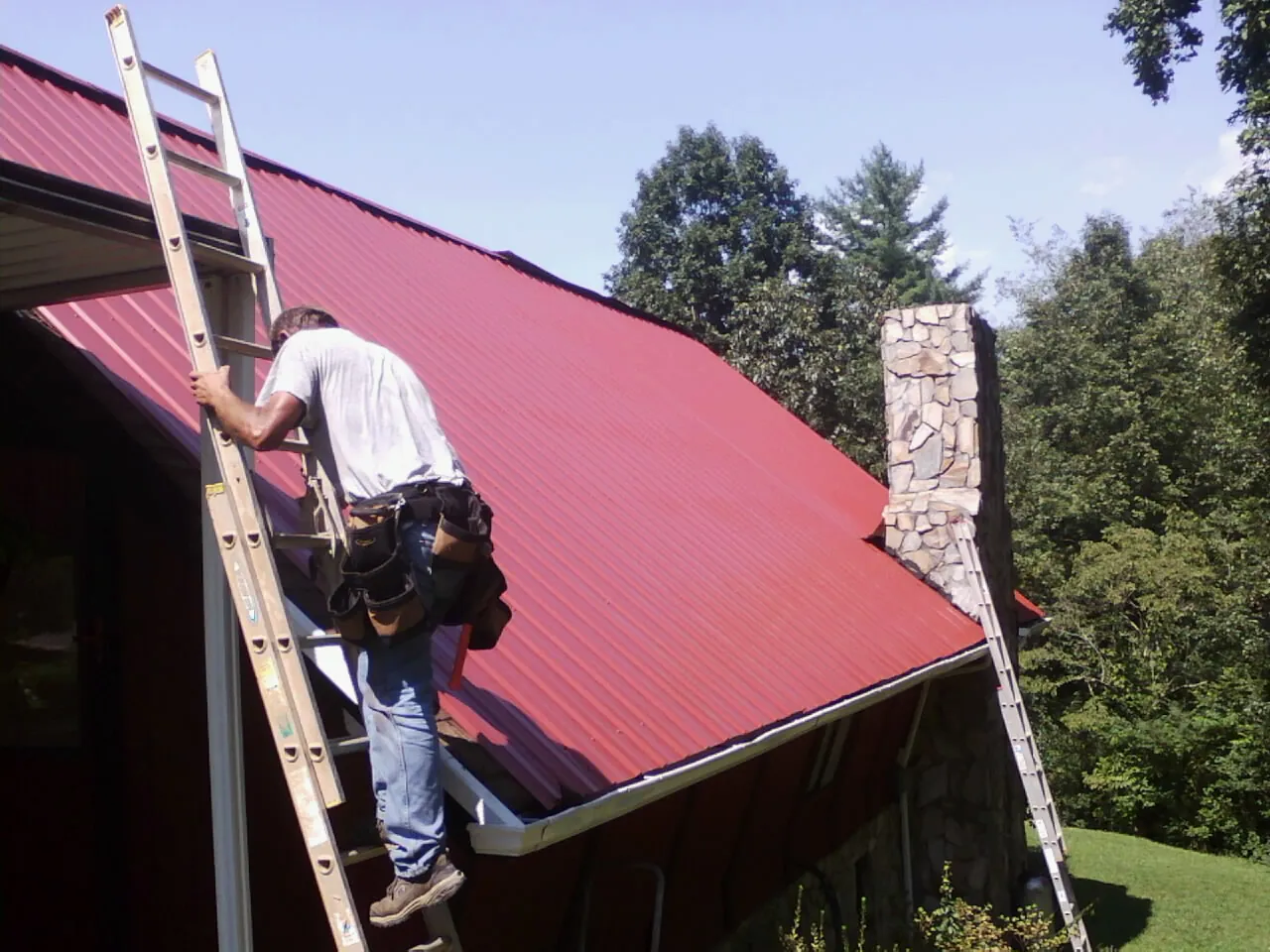 Roofer on a ladder beside a red metal roof and stone chimney — Soffit & Fascia Repair by Roofing Brothers in West Creek
